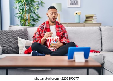 Young hispanic man eating popcorn watching movie on tablet device afraid and shocked with surprise and amazed expression, fear and excited face.  - Powered by Shutterstock