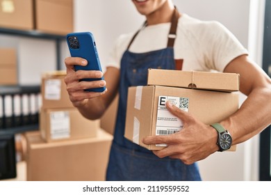 Young Hispanic Man Ecommerce Business Worker Holding Packages And Using Smartphone At Office