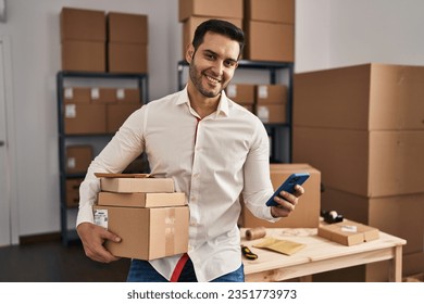 Young hispanic man e-commerce business worker using smartphone holding packages at office - Powered by Shutterstock