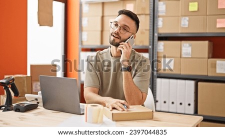Young hispanic man ecommerce business worker talking on smartphone writing address on package at office