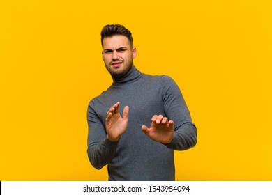 Young Hispanic Man Feeling Disgusted And Nauseous, Backing Away From Something Nasty, Smelly Or Stinky, Saying Yuck Against Orange Wall