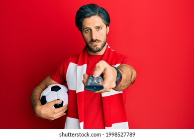 Young Hispanic Man Football Hooligan Holding Ball Using Tv Control Depressed And Worry For Distress, Crying Angry And Afraid. Sad Expression. 