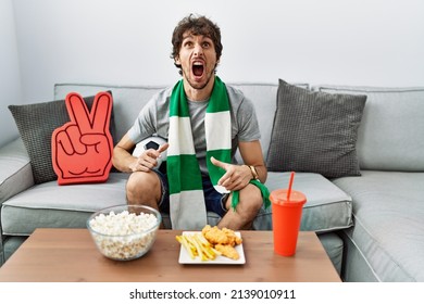 Young hispanic man football hooligan cheering game at home angry and mad screaming frustrated and furious, shouting with anger. rage and aggressive concept.  - Shutterstock ID 2139010911