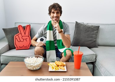 Young Hispanic Man Football Hooligan Cheering Game At Home With Hand On Chin Thinking About Question, Pensive Expression. Smiling And Thoughtful Face. Doubt Concept. 