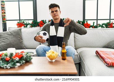 Young hispanic man football hooligan holding ball and beer with angry face, negative sign showing dislike with thumbs down, rejection concept 