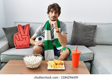 Young Hispanic Man Football Hooligan Cheering Game At Home With Hand On Chin Thinking About Question, Pensive Expression. Smiling With Thoughtful Face. Doubt Concept. 