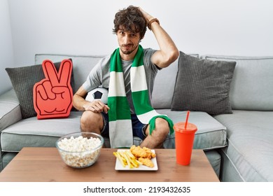 Young Hispanic Man Football Hooligan Cheering Game At Home Confuse And Wondering About Question. Uncertain With Doubt, Thinking With Hand On Head. Pensive Concept. 