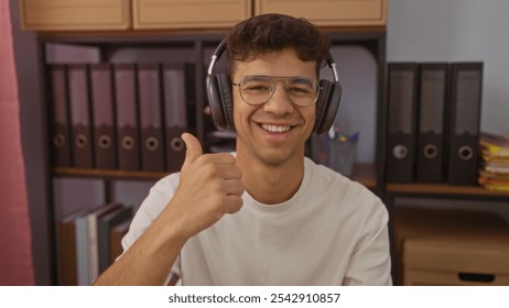 Young hispanic man giving a thumbs up while wearing headphones and smiling, seated in an office with shelves of files in the background. - Powered by Shutterstock