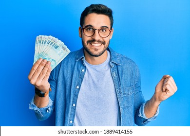 Young Hispanic Man Holding 100 Brazilian Real Banknotes Screaming Proud, Celebrating Victory And Success Very Excited With Raised Arm 