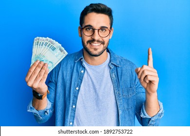 Young hispanic man holding 100 brazilian real banknotes smiling with an idea or question pointing finger with happy face, number one 