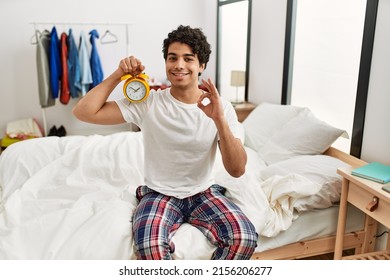 Young hispanic man holding alarm clock sitting on the bedroom doing ok sign with fingers, smiling friendly gesturing excellent symbol 