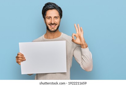 Young hispanic man holding blank empty banner doing ok sign with fingers, smiling friendly gesturing excellent symbol  - Shutterstock ID 1922211659