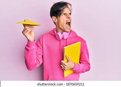 Young Hispanic Man Holding Book And Paper Airplane Angry And Mad Screaming Frustrated And Furious, Shouting With Anger. Rage And Aggressive Concept. 