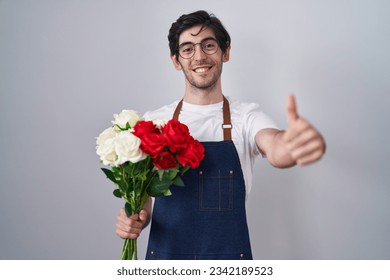 Young hispanic man holding bouquet of white and red roses approving doing positive gesture with hand, thumbs up smiling and happy for success. winner gesture.  - Powered by Shutterstock