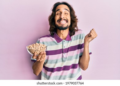 Young Hispanic Man Holding Chickpeas Bowl Screaming Proud, Celebrating Victory And Success Very Excited With Raised Arm 