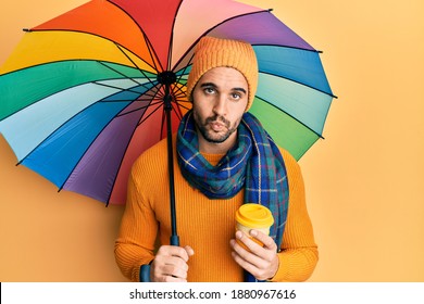 Young Hispanic Man Holding Colorful Umbrella Drinking Take Away Coffee Looking At The Camera Blowing A Kiss Being Lovely And Sexy. Love Expression. 