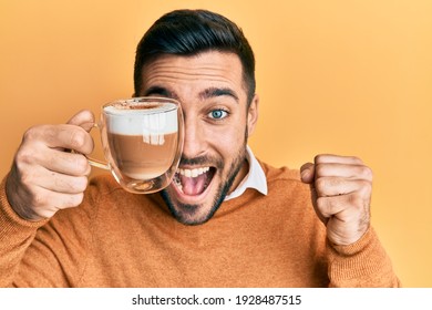 Young Hispanic Man Holding Cup Of Coffee Over Eye Screaming Proud, Celebrating Victory And Success Very Excited With Raised Arm 