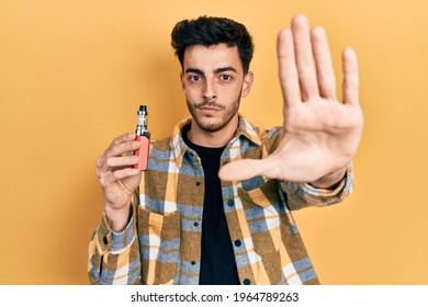 Young hispanic man holding electronic cigarette with open hand doing stop sign with serious and confident expression, defense gesture  - Powered by Shutterstock