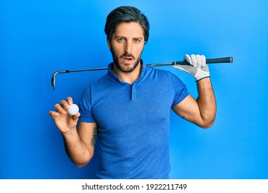 Young Hispanic Man Holding Golf Ball In Shock Face, Looking Skeptical And Sarcastic, Surprised With Open Mouth 