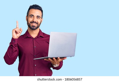 Young Hispanic Man Holding Laptop Surprised With An Idea Or Question Pointing Finger With Happy Face, Number One 