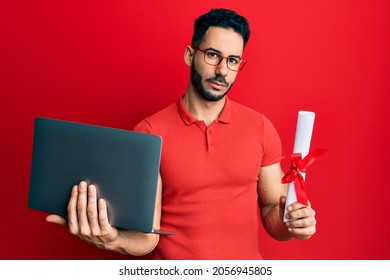 Young Hispanic Man Holding Laptop And Diploma Relaxed With Serious Expression On Face. Simple And Natural Looking At The Camera. 