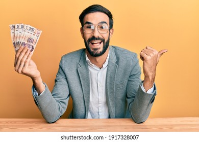 Young hispanic man holding mexican pesos sitting on the table pointing thumb up to the side smiling happy with open mouth 