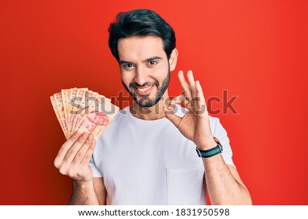 Young hispanic man holding mexican pesos doing ok sign with fingers, smiling friendly gesturing excellent symbol 