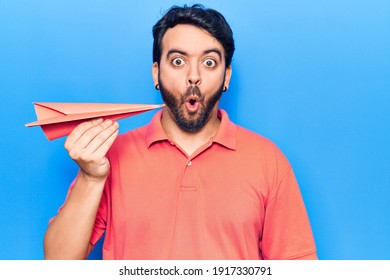 Young Hispanic Man Holding Paper Airplane Scared And Amazed With Open Mouth For Surprise, Disbelief Face 