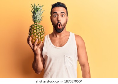 Young Hispanic Man Holding Pineapple Scared And Amazed With Open Mouth For Surprise, Disbelief Face 