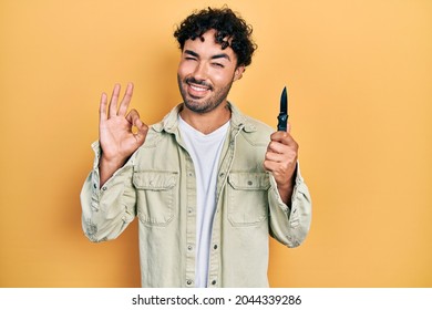 Young hispanic man holding pocket knife doing ok sign with fingers, smiling friendly gesturing excellent symbol 