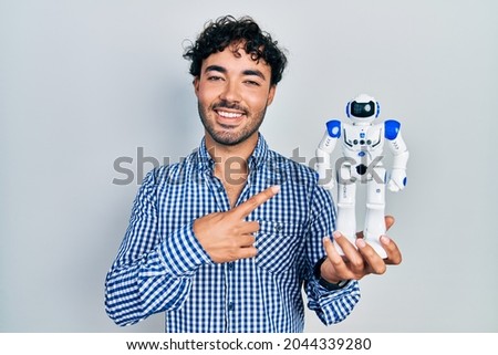 Young hispanic man holding robot toy smiling happy pointing with hand and finger 