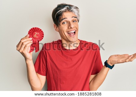 Young hispanic man holding second place badge celebrating achievement with happy smile and winner expression with raised hand 