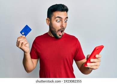 Young Hispanic Man Holding Smartphone And Credit Card Afraid And Shocked With Surprise And Amazed Expression, Fear And Excited Face. 