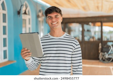 Young hispanic man holding a tablet at outdoors smiling a lot - Powered by Shutterstock