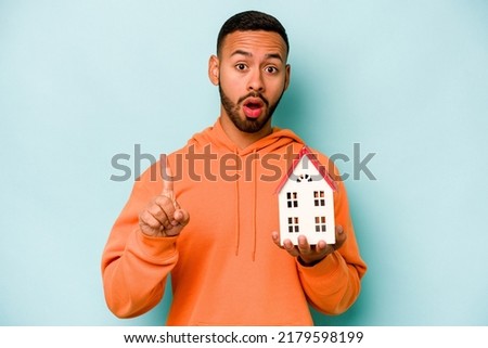 Young hispanic man holding a toy house isolated on blue background having some great idea, concept of creativity.