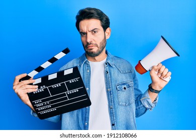 Young Hispanic Man Holding Video Film Clapboard And Megaphone Clueless And Confused Expression. Doubt Concept. 