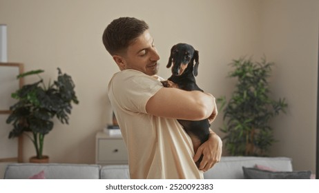 A young hispanic man holds his dachshund dog lovingly while standing in a cozy, well-decorated living room.