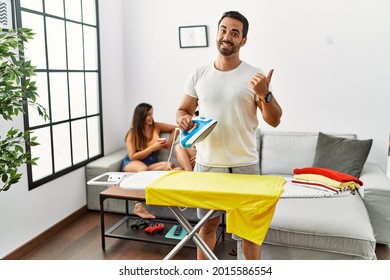 Young hispanic man ironing clothes at home pointing to the back behind with hand and thumbs up, smiling confident  - Powered by Shutterstock