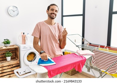 Young Hispanic Man Ironing Clothes At Home Doing Money Gesture With Hands, Asking For Salary Payment, Millionaire Business 