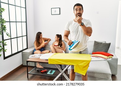 Young Hispanic Man Ironing Clothes At Home With Hand On Chin Thinking About Question, Pensive Expression. Smiling And Thoughtful Face. Doubt Concept. 