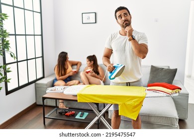 Young Hispanic Man Ironing Clothes At Home With Hand On Chin Thinking About Question, Pensive Expression. Smiling With Thoughtful Face. Doubt Concept. 