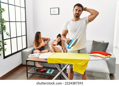 Young Hispanic Man Ironing Clothes At Home Confuse And Wondering About Question. Uncertain With Doubt, Thinking With Hand On Head. Pensive Concept. 