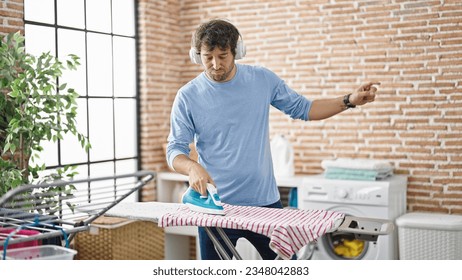 Young hispanic man listening to music ironing clothes dancing at laundry room - Powered by Shutterstock