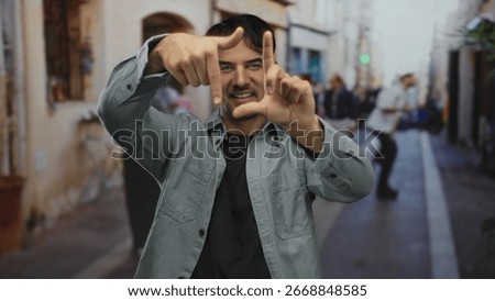 Young hispanic man making framing gesture with hands on a bustling city street outdoor showcasing creativity in photography in an urban environment bustling with activity.