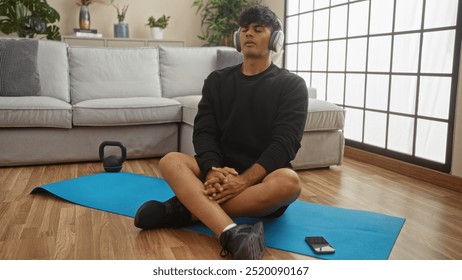 Young hispanic man meditating on a blue yoga mat wearing headphones in the living room of his apartment. - Powered by Shutterstock