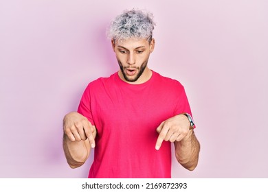 Young Hispanic Man With Modern Dyed Hair Wearing Casual Pink T Shirt Pointing Down With Fingers Showing Advertisement, Surprised Face And Open Mouth 