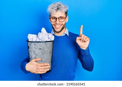 Young Hispanic Man With Modern Dyed Hair Holding Paper Bin Full Of Crumpled Papers Surprised With An Idea Or Question Pointing Finger With Happy Face, Number One 