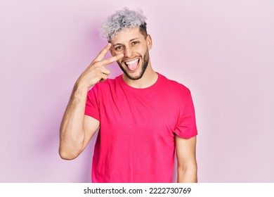 Young hispanic man with modern dyed hair wearing casual pink t shirt doing peace symbol with fingers over face, smiling cheerful showing victory 