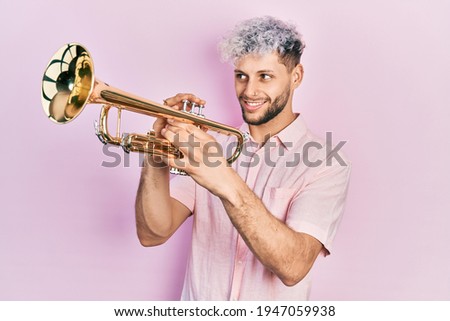 Young hispanic man with modern dyed hair playing trumpet smiling looking to the side and staring away thinking. 