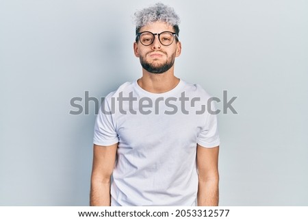 Young hispanic man with modern dyed hair wearing white t shirt and glasses puffing cheeks with funny face. mouth inflated with air